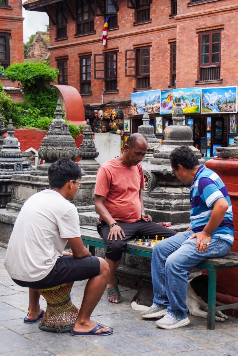 Three men playing chess on a bench in front of stone stupas in Patan